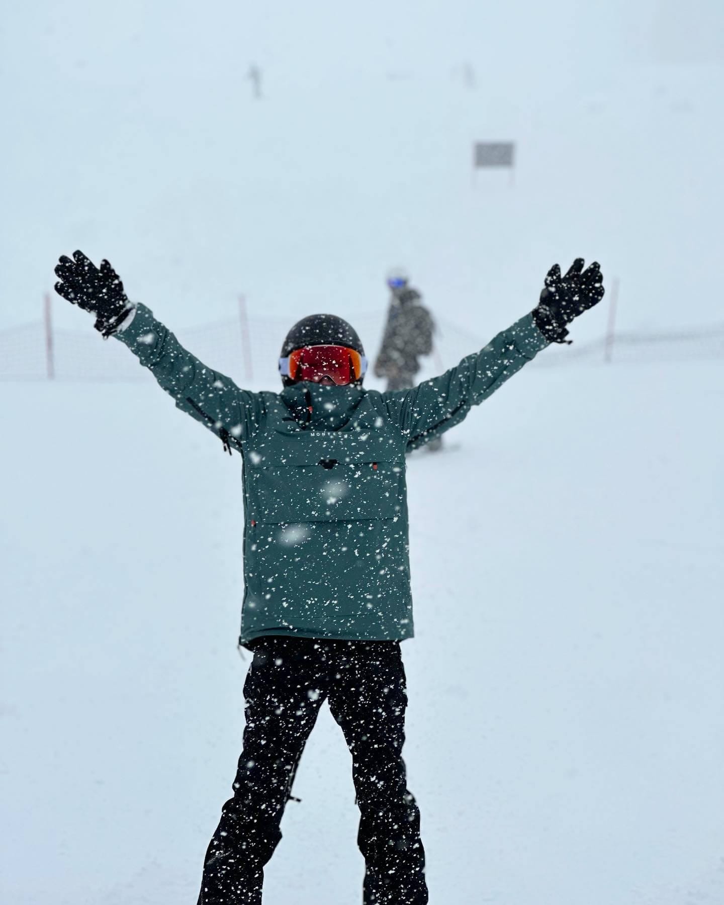 Jenna Felkey, wearing a snowboarding helmet, teal jacket, and black pants, poses with her arms up on snowy hill.