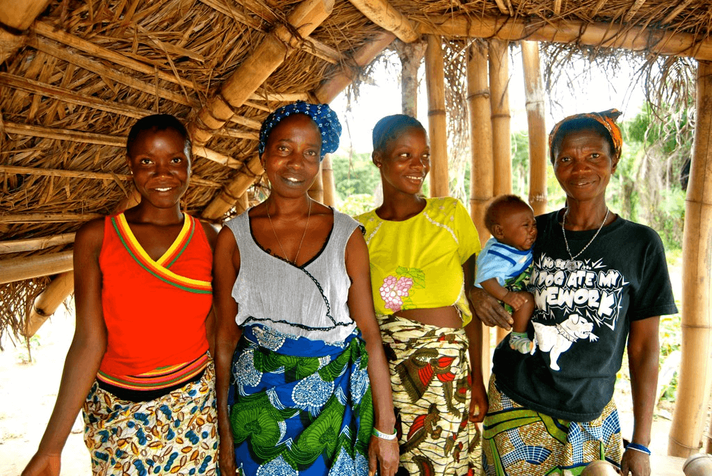 Four women, two expectant mothers and two midwives, stand for a photo. The woman on the right holds a young baby. 