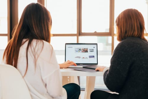 Two women sitting in front of a laptop at a desk.