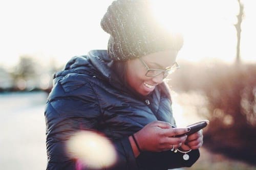 Woman of color with a dark winter jacket and hat holding a cell phone.